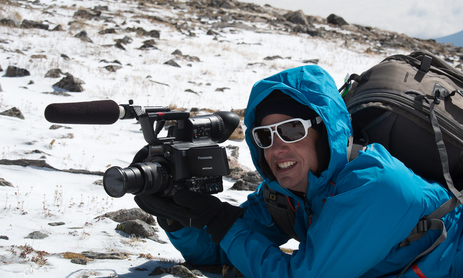 Man in a blue overcoat with hood up, laying on snow, holding a video camera.