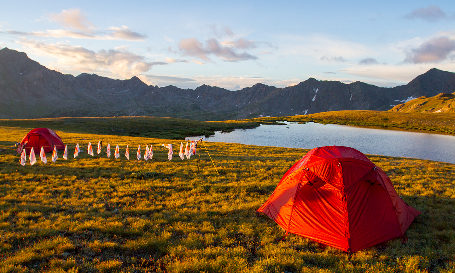 basecamp image showing two orange tents and a sting of flags. mountains and a lake are in the background