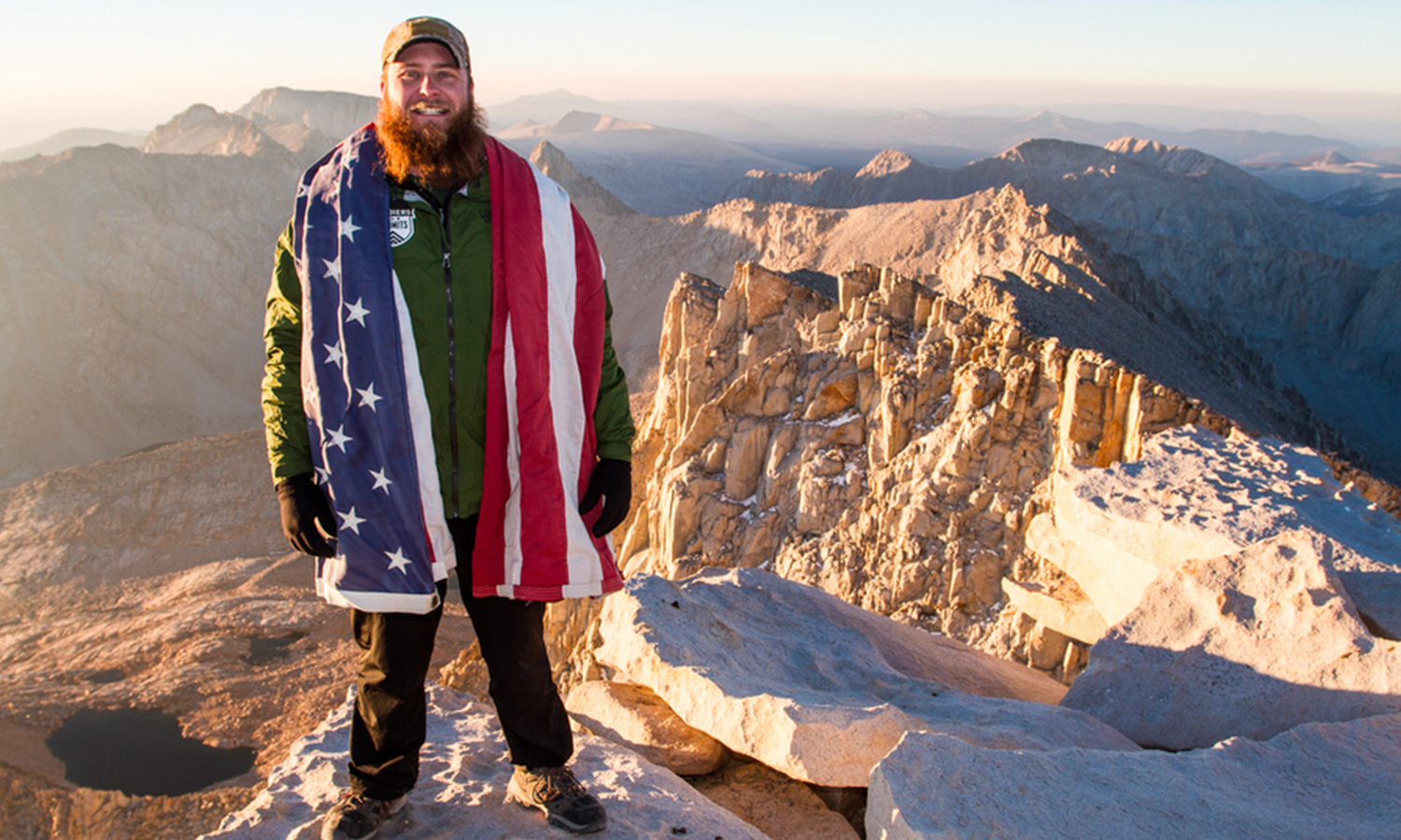 man stands a top mountain with american flag draped over his arms